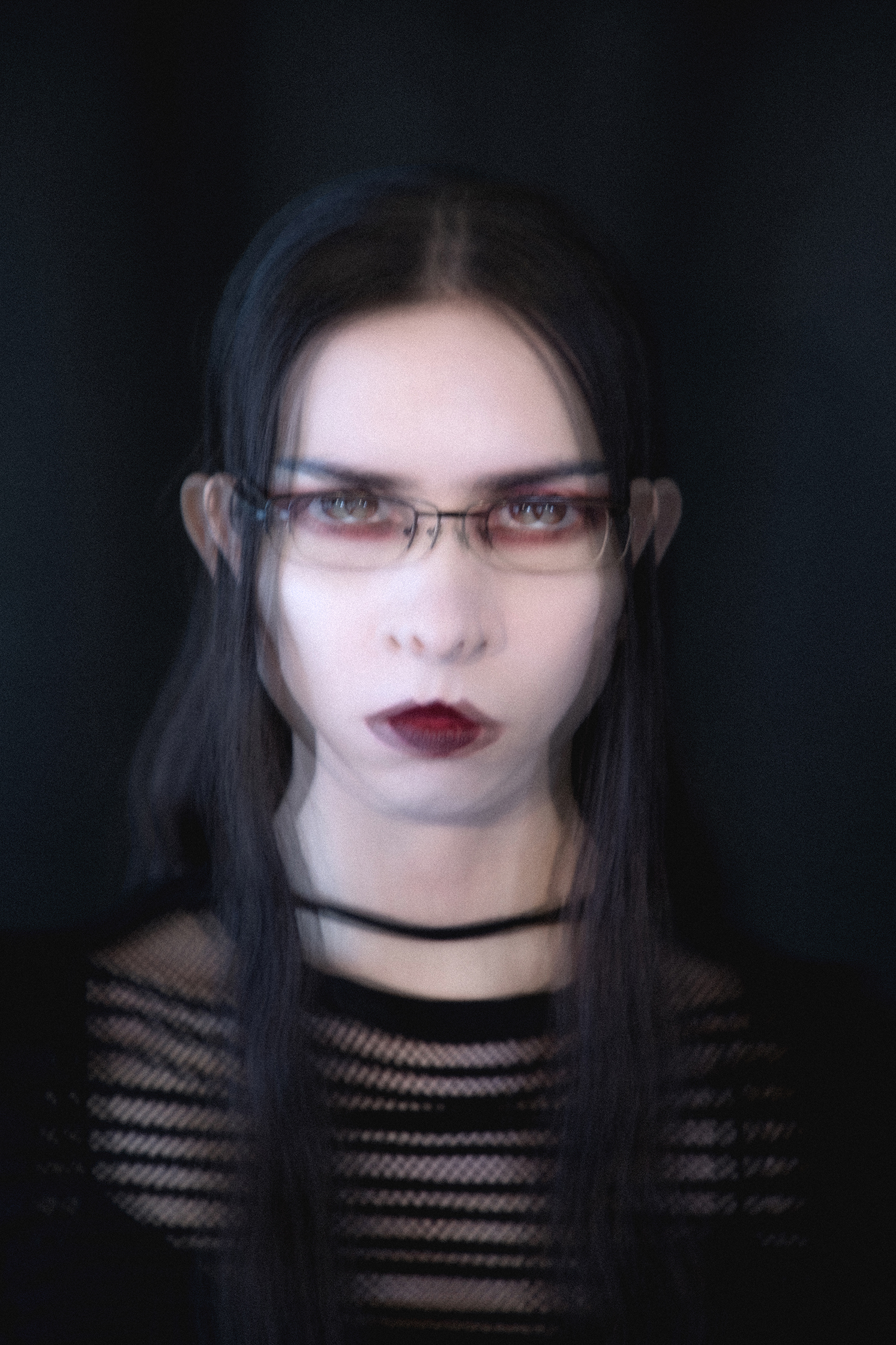 Blurry image of a female model with pale skin and glasses on with dark eye makeup looking into the camera
